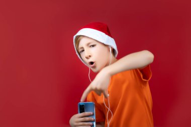 Kırmızı arka planda bir çocuk ve Noel Baba 'nın Noel şapkası telefon tutarken yüzünü buruşturuyor ve müzik dinliyor.