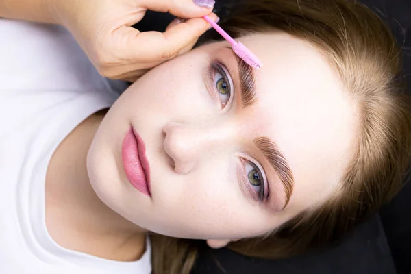 the face of a beautiful blonde model after the eyebrow lamination procedure, the master combs the eyebrows with a brush