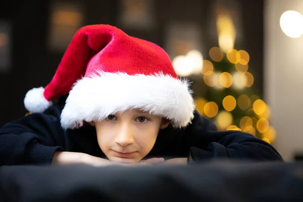 サンタの帽子の男の子がソファの上であごを折りクリスマスツリーのおもちゃを背景にカメラを見て彼は少し悲しい — ストック写真