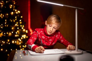 Noel ağacının arka planında kırmızı bir Noel süveteri giyen bir çocuk bir odada bir masada oturuyor.