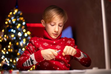 Kırmızı Noel süveteri giyen bir çocuk beyaz bir masada bir lambanın önüne çiziyor.