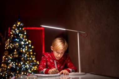 Noel ağacının arka planında kırmızı bir Noel süveteri giyen bir çocuk odasında oturuyor ve resim çiziyor.