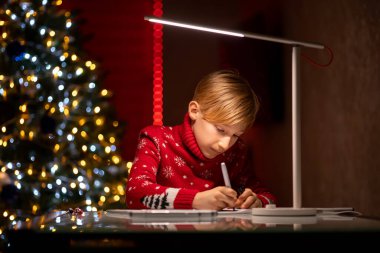 Noel ağacının arka planında kırmızı bir Noel süveteri giyen bir çocuk odasında oturuyor ve resim çiziyor.