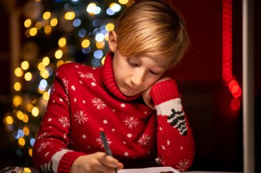 Noel ağacının arka planında kırmızı bir Noel süveteri giyen bir çocuk elini yüzüne bastırdı ve düşünceli bir şekilde çizdi.