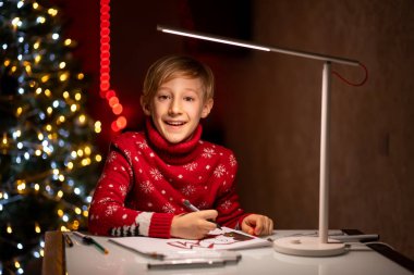 Noel ağacının arka planında kırmızı bir Noel süveteri giyen bir çocuk elinde kalemlerle gülümsüyor.