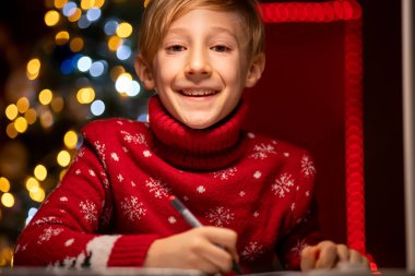 Noel ağacının arka planında kırmızı bir Noel süveteri giyen bir çocuk elinde kalemlerle gülümsüyor.