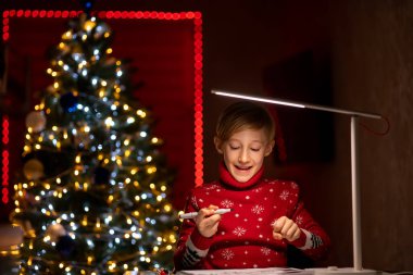 Noel ağacının arka planında kırmızı Noel süveteri giyen bir çocuk çizimlerini bir kalemle süslemeye hazırlanıyor.
