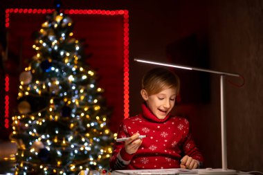 Noel ağacının arka planında kırmızı Noel süveteri giyen bir çocuk çizimlerini bir kalemle süslemeye hazırlanıyor.