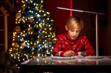 Noel ağacının arka planında kırmızı Noel süveteri giyen bir çocuk çizimlerini bir kalemle süslemeye hazırlanıyor.