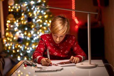 Noel ağacının arka planında kırmızı bir Noel süveteri giyen bir çocuk çizimin üzerine eğilip onu bir kalemle süsledi.