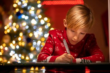 Kırmızı Noel süveteri giymiş bir çocuk Noel ağacının arka planında keçeli kalemle çizim yapmaya eğilmiş.