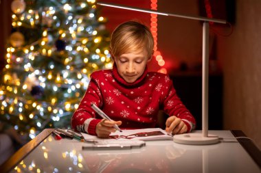 Kırmızı Noel süveteri giymiş bir çocuk bir lambayla aydınlatılmış bir Noel ağacının arka planında elinde bir çizim kalemi tutuyor.