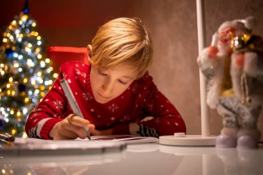 Noel ağacının arka planında kırmızı süveter giyen bir çocuk yeni yıl için çizimini bitirir.