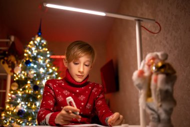 Noel ağacının arka planında kırmızı süveter giyen bir çocuk yeni yıl için çizimini bitirir.