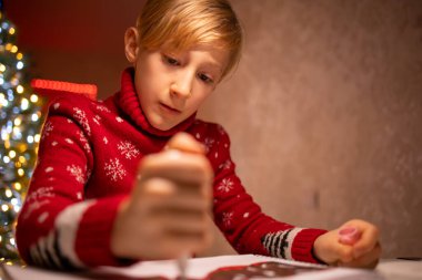 Noel ağacının arka planında kırmızı Noel süveteri giyen bir çocuk yeni yılın tamamlanmış çizimine bakar.