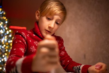 Noel ağacının arka planında kırmızı Noel süveteri giyen bir çocuk yeni yılın tamamlanmış çizimine bakar.