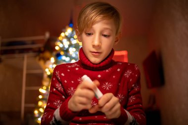 Noel ağacının arka planında kırmızı Noel süveteri giyen bir çocuk önünde bir kalem tutuyor ve onu açmak istiyor.