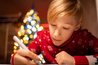 Noel ağacının arka planında kırmızı bir Noel süveteri giyen bir çocuk masaya eğilir ve bir resim çizmeye çalışır.