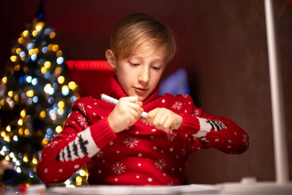 Kırmızı Noel süveteri giyen bir çocuk beyaz bir masada bir lambanın önüne çiziyor.