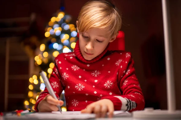 Noel ağacının arka planında kırmızı Noel süveteri giyen bir çocuk keçeli kalemle resim çiziyor.