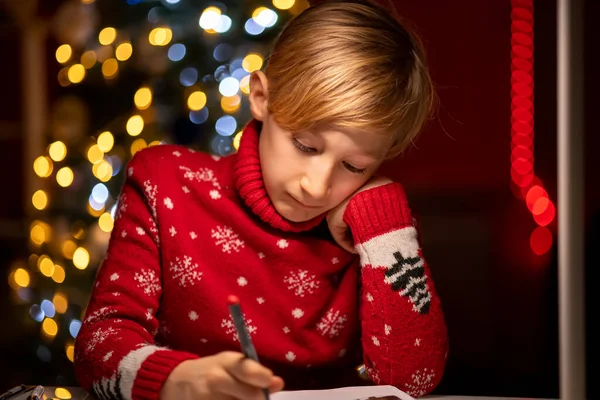 Noel ağacının arka planında kırmızı bir Noel süveteri giyen bir çocuk elini yüzüne bastırdı ve düşünceli bir şekilde çizdi.