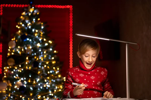 Noel ağacının arka planında kırmızı Noel süveteri giyen bir çocuk çizimlerini bir kalemle süslemeye hazırlanıyor.