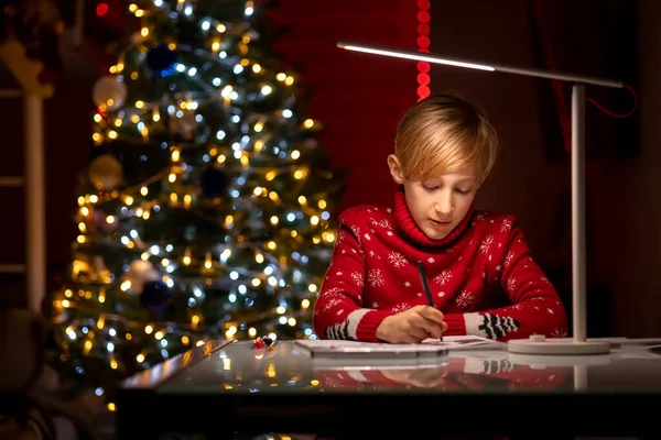 Noel ağacının arka planında kırmızı Noel süveteri giyen bir çocuk çizimlerini bir kalemle süslemeye hazırlanıyor.