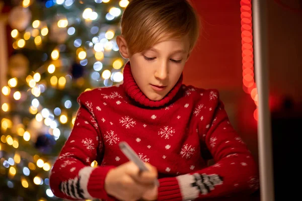 Kırmızı Noel süveteri giymiş bir çocuk Noel ağacının arka planında keçeli kalemle çizim yapmaya eğilmiş.
