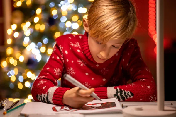 Kırmızı Noel süveteri giymiş bir çocuk Noel ağacının arka planında keçeli kalemle çizim yapmaya eğilmiş.