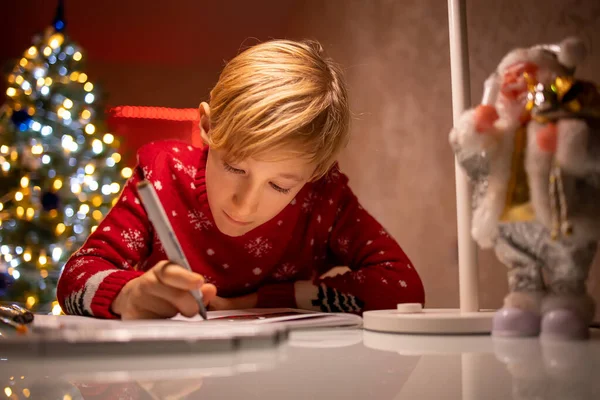 Noel ağacının arka planında kırmızı süveter giyen bir çocuk yeni yıl için çizimini bitirir.