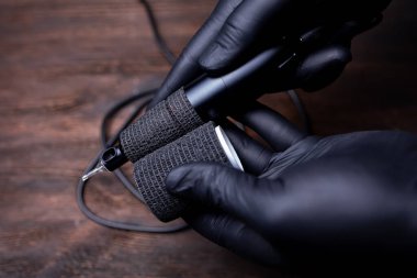 Siyah steril eldivenli bir makyaj sanatçısının elleri siyah bir bandaj bandıyla sarılmış dövme makinesini koruyor.