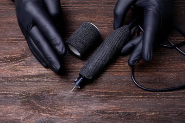 Siyah steril eldivenli bir makyaj sanatçısının elleri önünde tahta bir masaya sarılı bir dövme makinesi yatıyor.