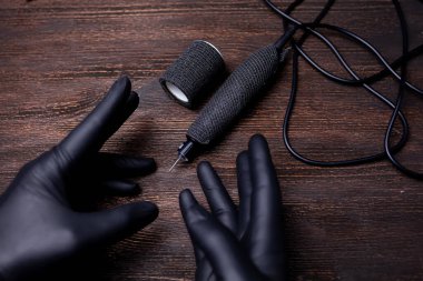 Siyah steril eldivenli bir makyaj sanatçısının elleri siyah bir bandaj bandıyla sarılı bir dövme makinesini işaret ediyor.