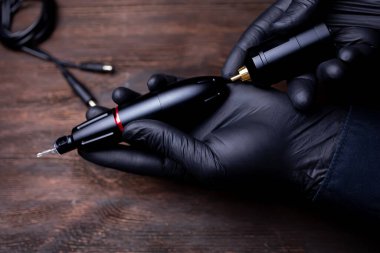 Siyah steril eldivenli makyajcının elleri dövme makinesinin kablosuz aküsünü bağlamaya çalışıyor.