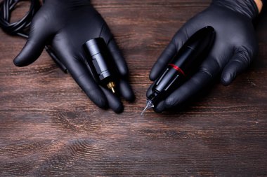 Avuçlarında dövme makinesi ve bu makine için kablosuz cihaz olan siyah steril eldivenli bir makyaj sanatçısının elleri.