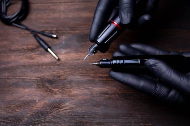 Siyah steril eldivenli bir makyaj sanatçısının elleri iki dövme makinesini elinde tutuyor ve birbirlerine kartuşlar yolluyorlar.