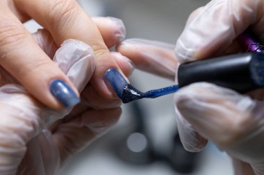 Manikür servisinin ustası, mavi jel cilası sürdüğü yakın plan manikür yaptırıyor.