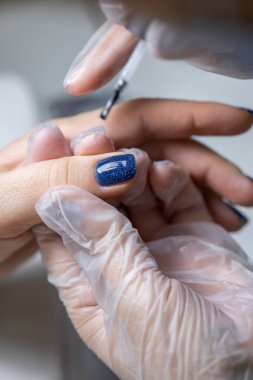 Manikür ustası müşterinin parmağını tutar ve tırnak cilasına mavi jöle sürer.
