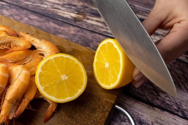 on a wooden stand, a lemon is cut with a knife near boiled shrimp