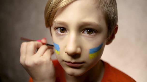 戦争に反対する子供 頬にウクライナの旗が描かれた少年がカメラに向かってじっと見つめている — ストック動画