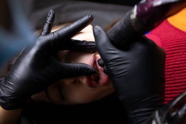 Siyah steril eldivenli bir dövme sanatçısı dövme makinesini tutuyor ve modelin alt dudağına dövme yaptırıyor.