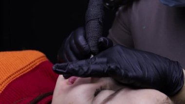 Modelin üzerine eğilen usta dövme makinesi ve kırmızı pigment kullanarak model için kalıcı dudak makyaj prosedürleri gerçekleştirir.