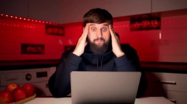 Siyah başlıklı sakallı bir adam dizüstü bilgisayarın önünde oturuyor. Elleriyle çeşitli jestler yapıyor ve kameraya bakıyor.