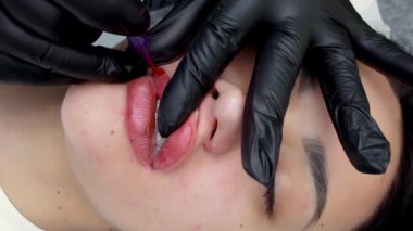 Siyah eldivenli kalıcı makyaj ustası bir dövme makinesi tutuyor ve esmer bir model için kalıcı dudak makyajı yapıyor.