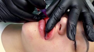 Bir dövme makinesi ve kırmızı pigmentle kalıcı dudak makyajı yapan model bir kızın dudaklarının yakın çekimi.