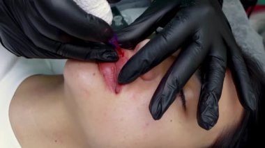 Siyah eldivenli kalıcı makyaj ustası bir dövme makinesi tutuyor ve esmer bir model için kalıcı dudak makyajı yapıyor.