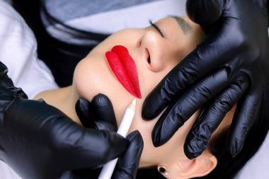 Siyah eldivenli dövme ustasının elleri beyaz bir kalem tutar ve kırmızı kalemle boyanmış dudakların dış hatlarını çizer.