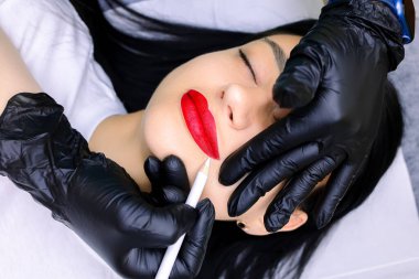 Siyah eldivenli dövme ustasının elleri beyaz bir kalem tutar ve kırmızı kalemle boyanmış dudakların dış hatlarını çizer.