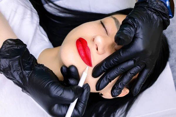 Siyah eldivenli dövme ustasının elleri beyaz bir kalem tutar ve kırmızı kalemle boyanmış dudakların dış hatlarını çizer.