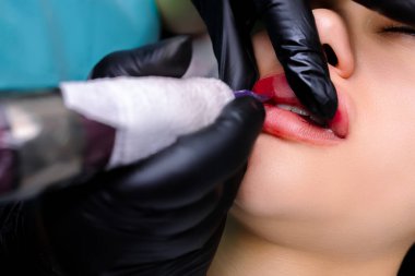 Dövmeci dudakları parmaklarıyla tutar, böylece dudakların alt kısmını kırmızı pigmentle tarar ve dövme makinesi kullanır.